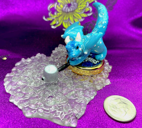 A small blue dragon smashes a sledgehammer down onto ice cubes from atop a mini-Icebreakers candy tin